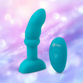 The B-Vibe Remote Rimming P-Spot Plug, a blue silicone vibrator with a curved tip and flared base, is displayed beside its oval remote against a soft purple and pink bokeh background.
